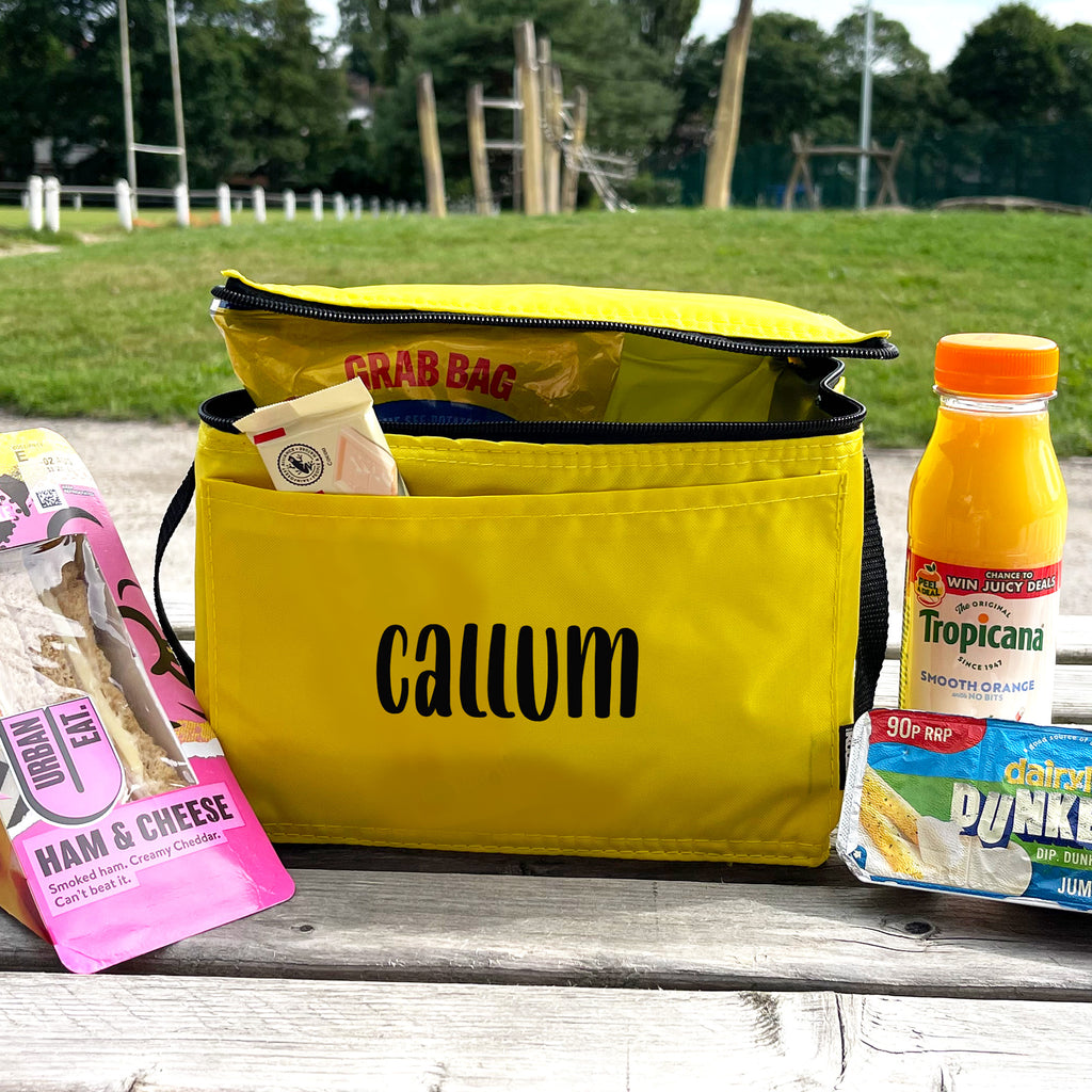 Yellow bag labeled 'Callum' with snacks and drinks on a bench outdoors