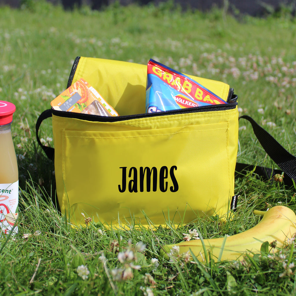 Yellow bag with 'James' printed on it, containing snacks and drinks, on a grassy background.
