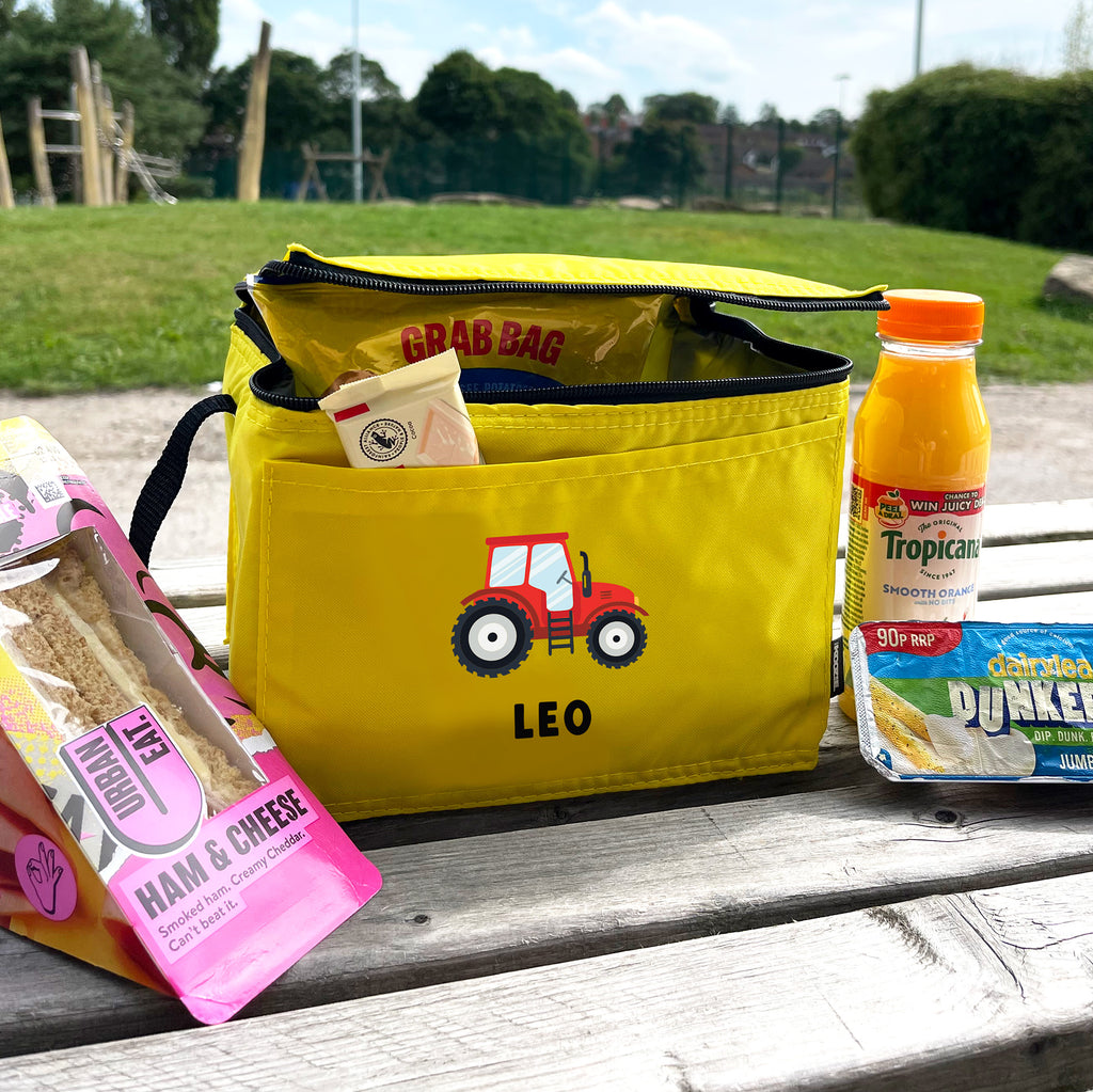 Yellow lunch bag with a red tractor design and 'Leo' text, containing snacks and a juice bottle on a wooden surface.