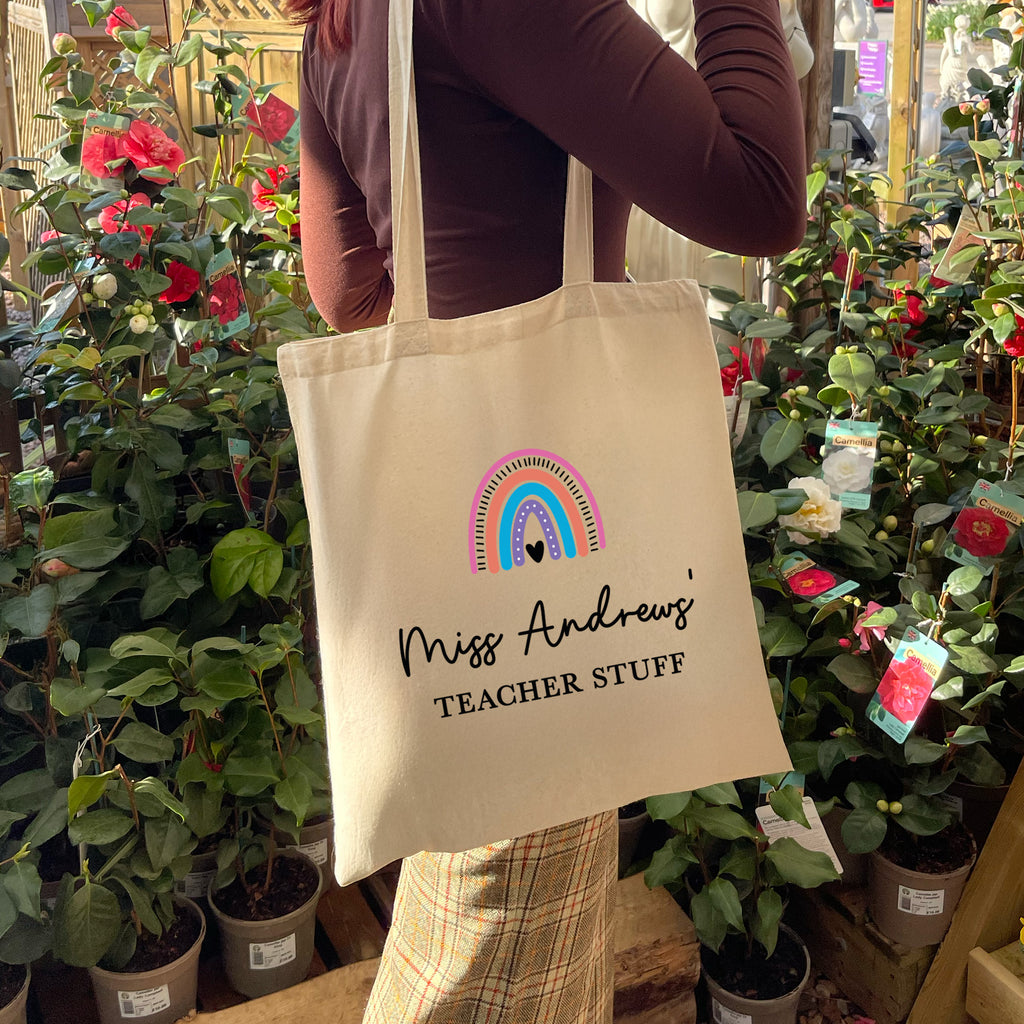 Person holding a tote bag with 'Miss Andrews Teacher Stuff' text and rainbow design among plants.