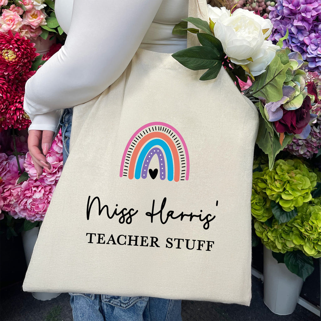 Tote bag with rainbow design and text 'Miss Harris' Teacher Stuff' held by a person surrounded by flowers.