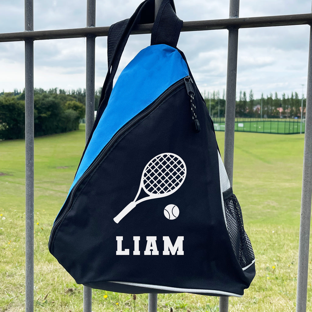 Black and blue sports bag with tennis racket and ball design, and 'LIAM' text, hanging on a metal fence with a grassy field background.