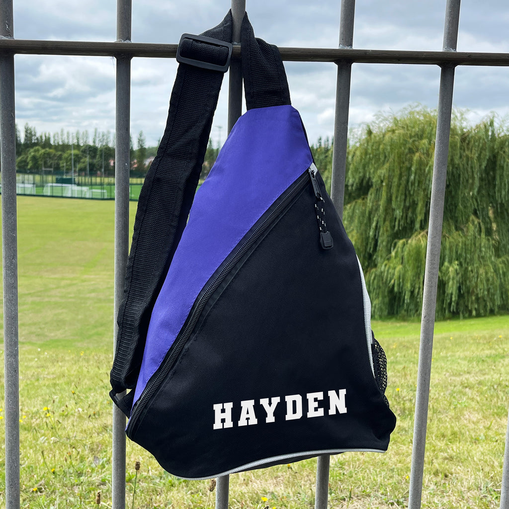 Black and purple sling bag with 'HAYDEN' printed on it, hanging on a metal fence with a grassy field and trees in the background.