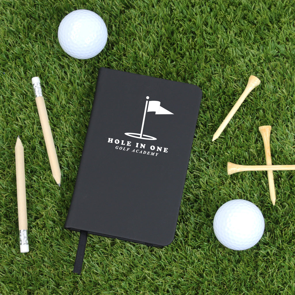 Black notebook with 'Hole in One Golf Academy' logo on grass with golf balls and tees.