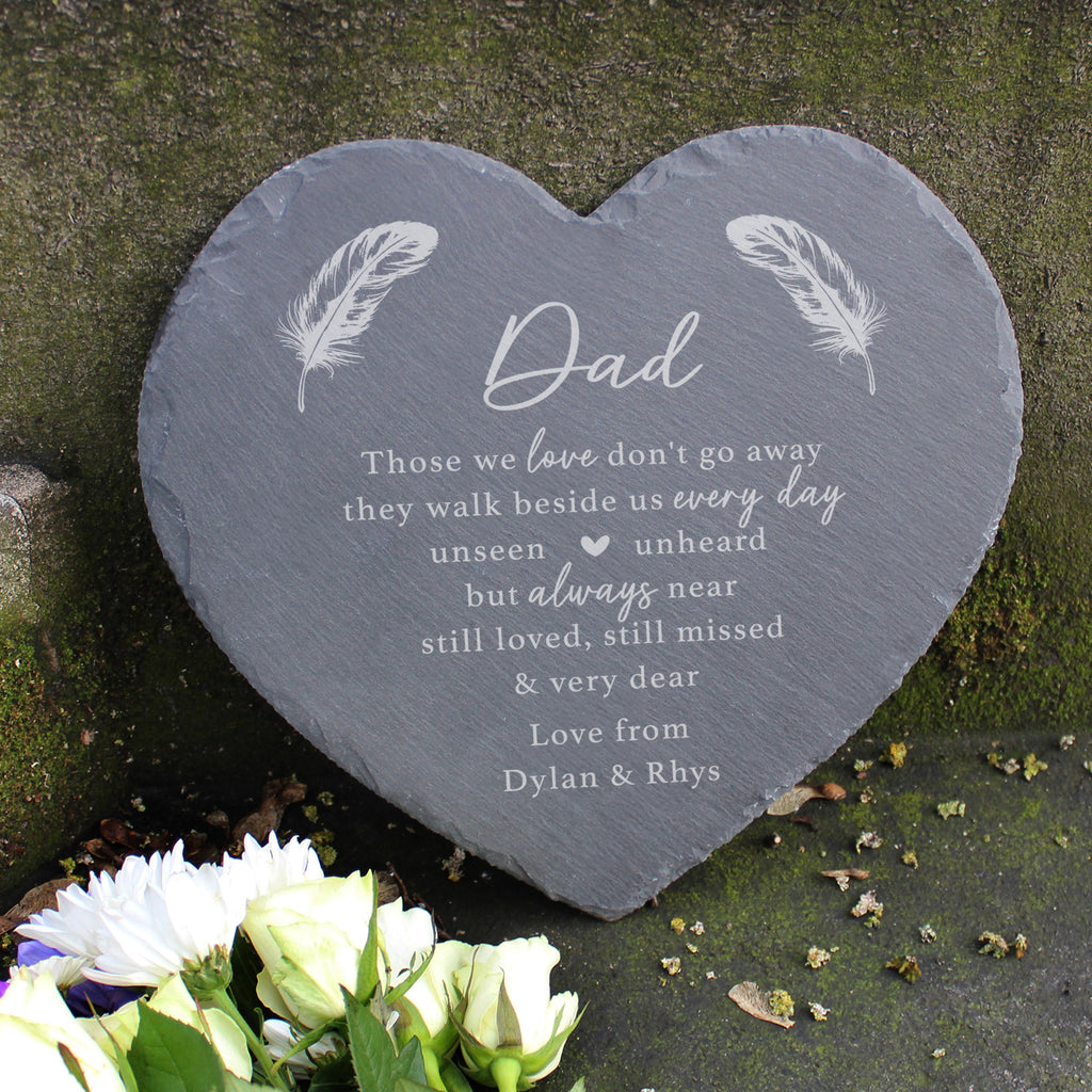 Heart-shaped slate plaque with engraved text and feathers on a stone surface with flowers.