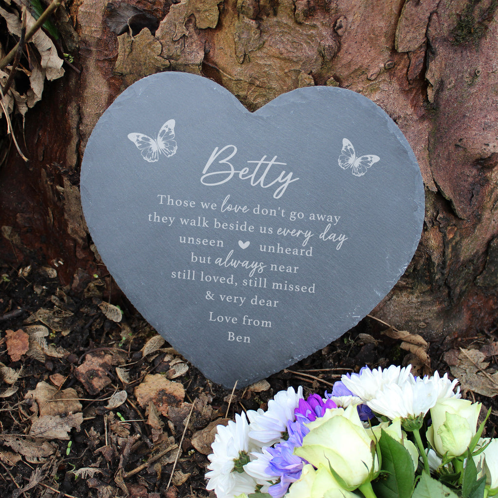 Heart-shaped memorial plaque with engraved text and butterflies on a tree trunk with flowers.