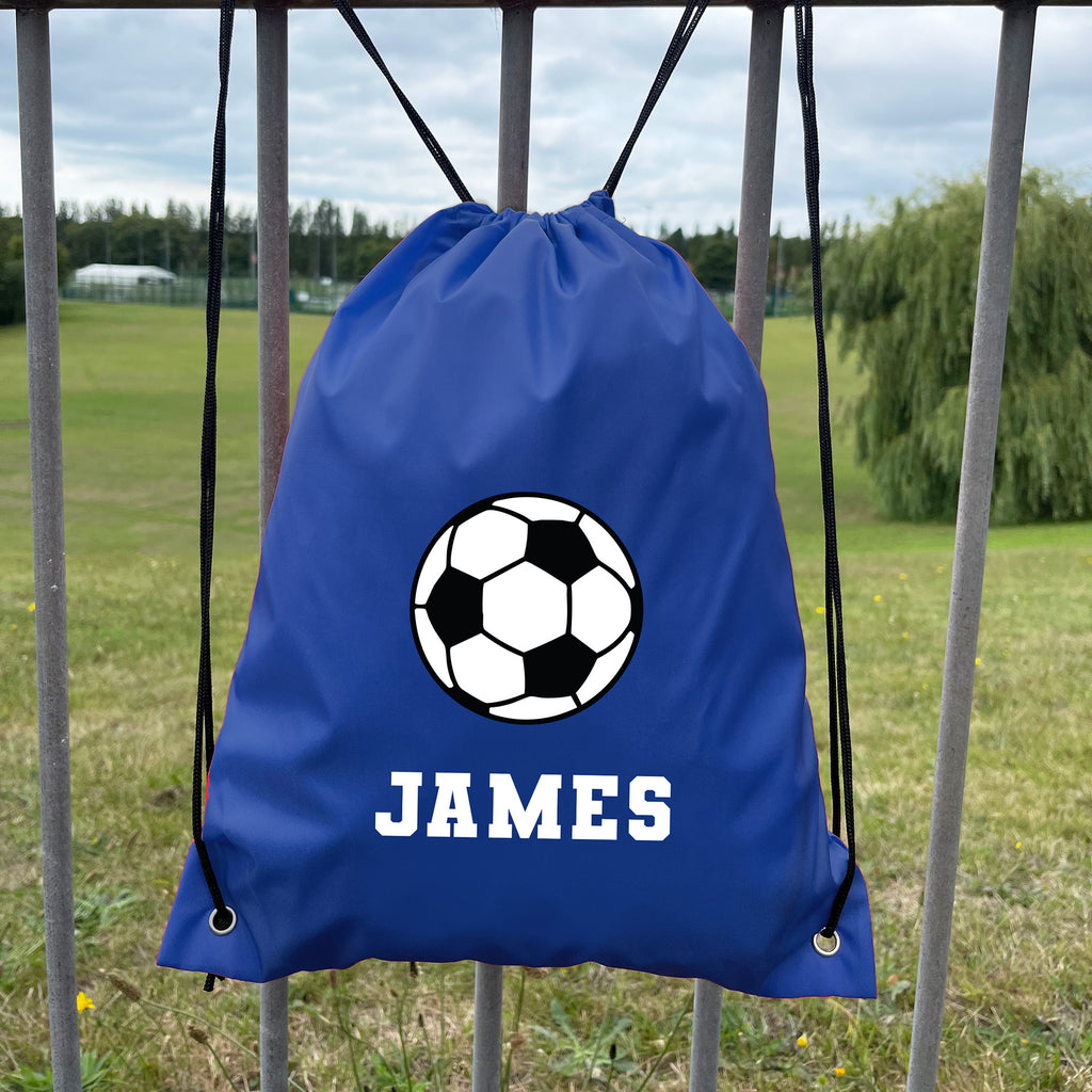 Blue drawstring bag with soccer ball design and 'JAMES' text, hanging on a metal fence with a grassy field background.