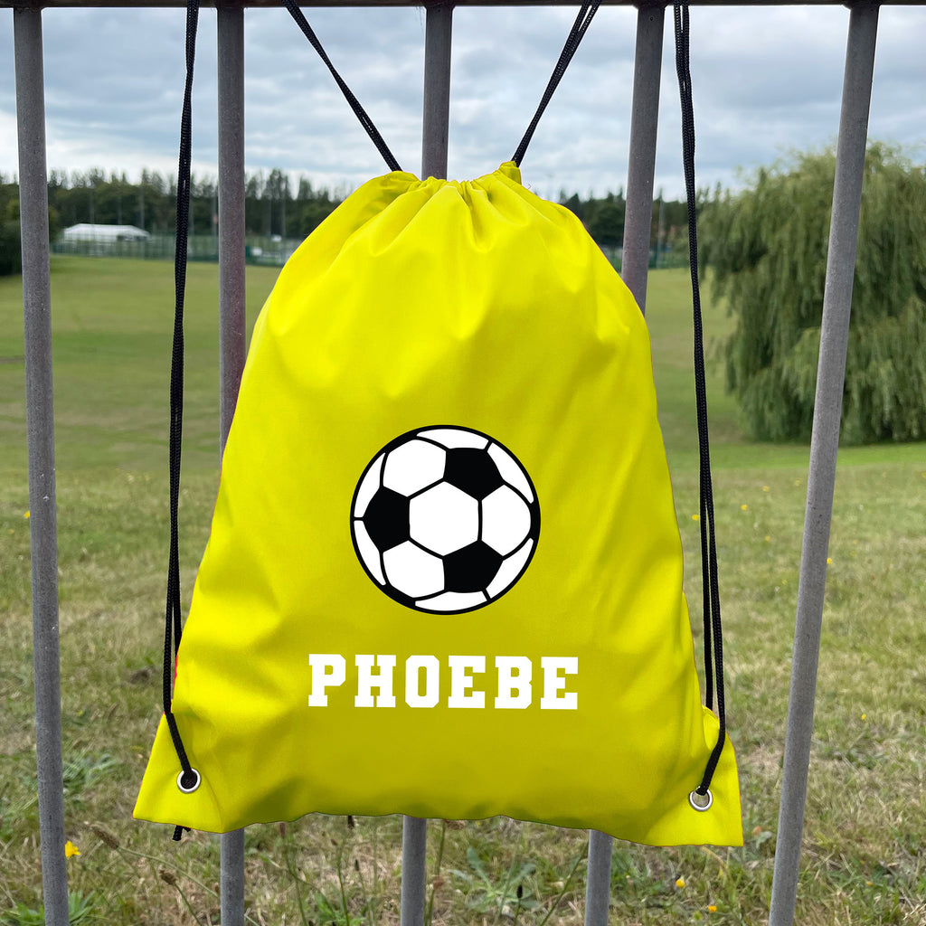 Yellow drawstring bag with soccer ball design and 'PHOEBE' text, hanging on a metal fence with a grassy field background.