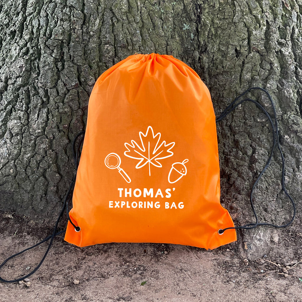 Orange drawstring bag labeled 'Thomas' Exploring Bag' against a tree trunk.