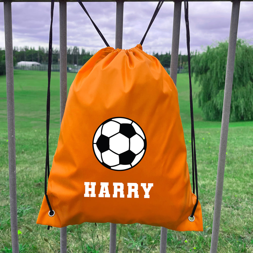 Orange drawstring bag with soccer ball design and 'HARRY' text, hanging on a metal fence with a grassy field background.