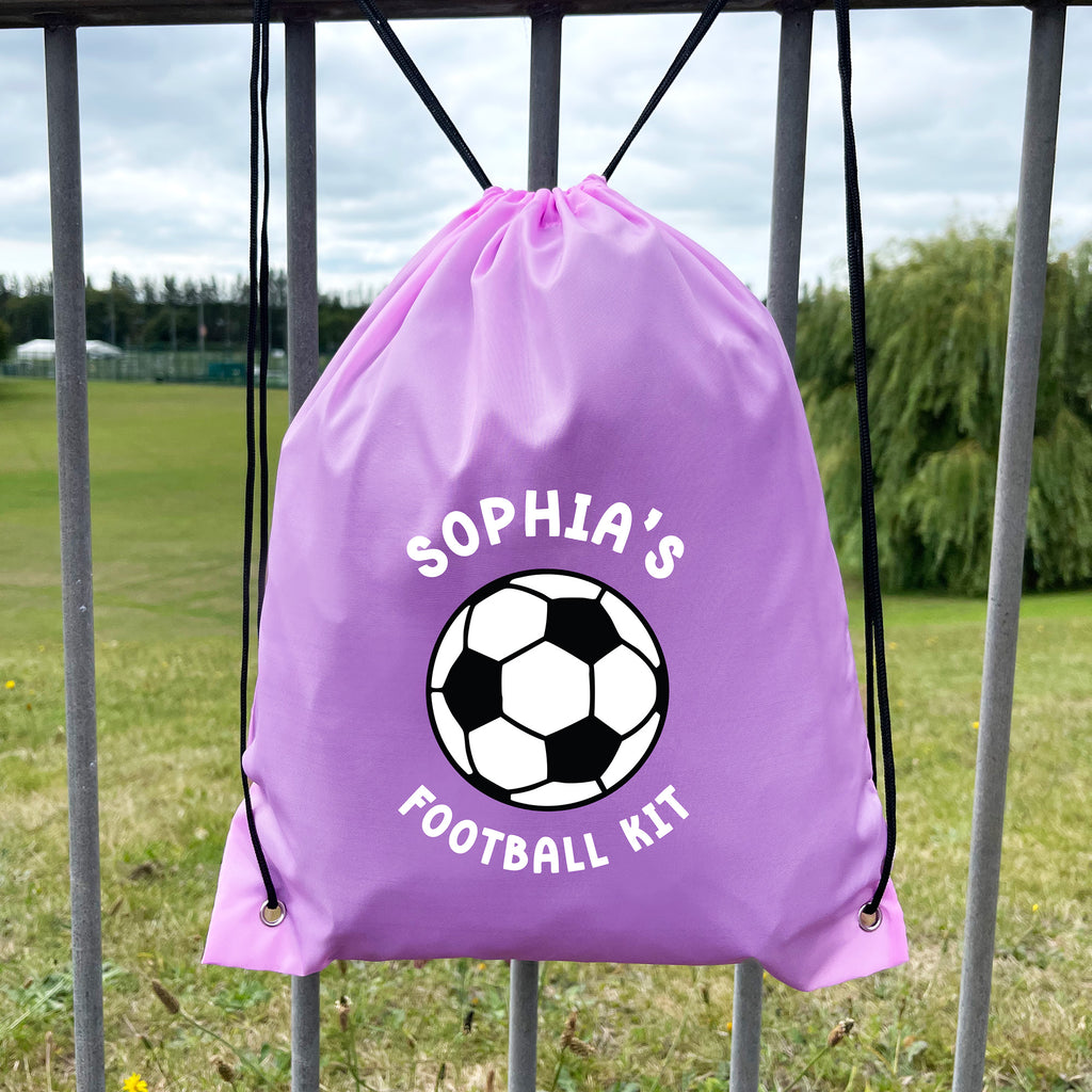 Purple drawstring bag with soccer ball design and 'Sophia's Football Kit' text, hanging on a metal fence with a grassy field background.
