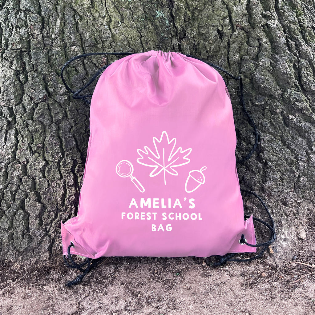 Pink drawstring bag with 'Amelia's Forest School Bag' text and graphics against a tree trunk.
