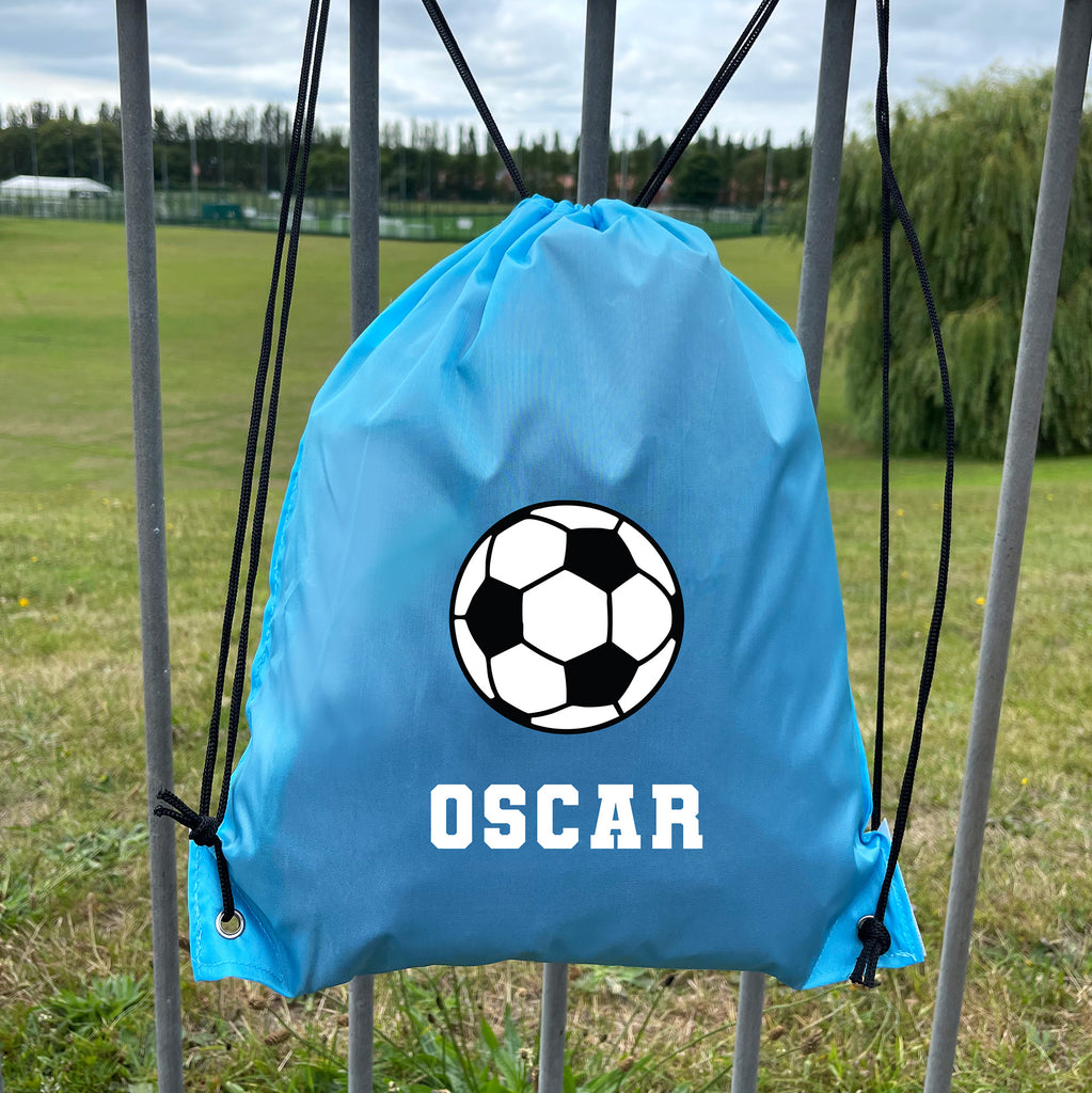 Blue drawstring bag with soccer ball design and 'OSCAR' text, hanging on a metal gate with a grassy field background.