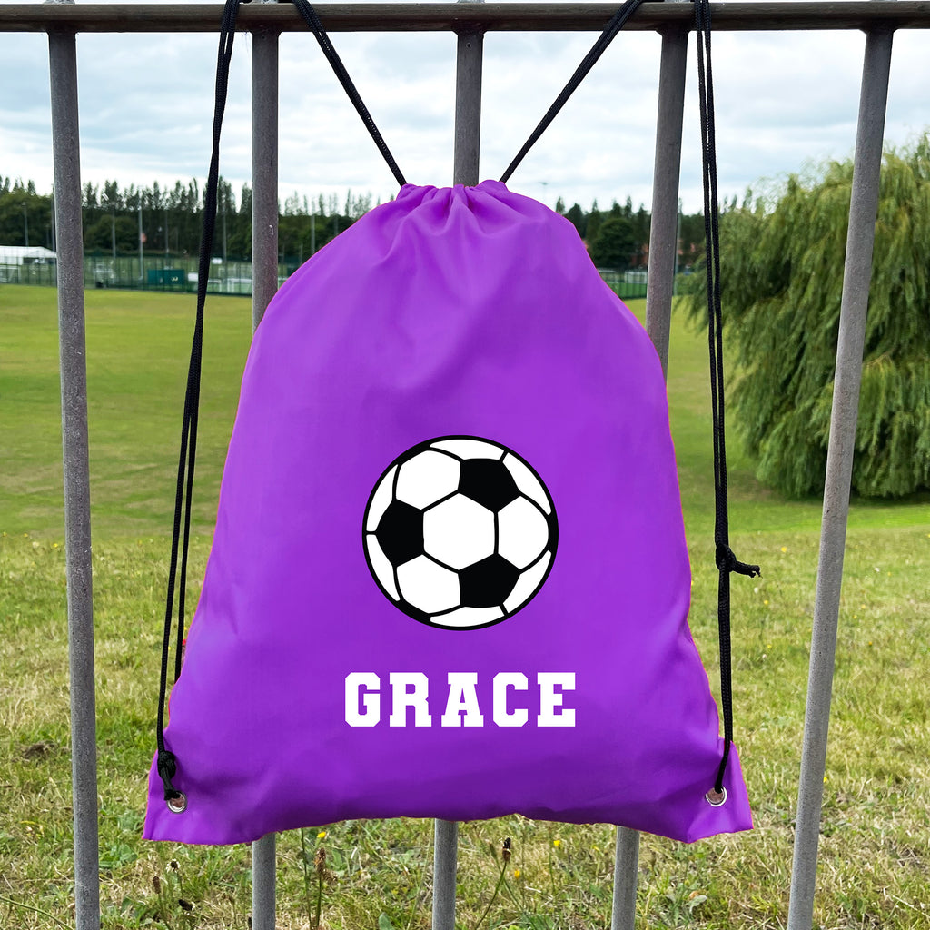 Purple drawstring bag with soccer ball design and 'GRACE' text, hanging on a metal fence with a grassy field background.
