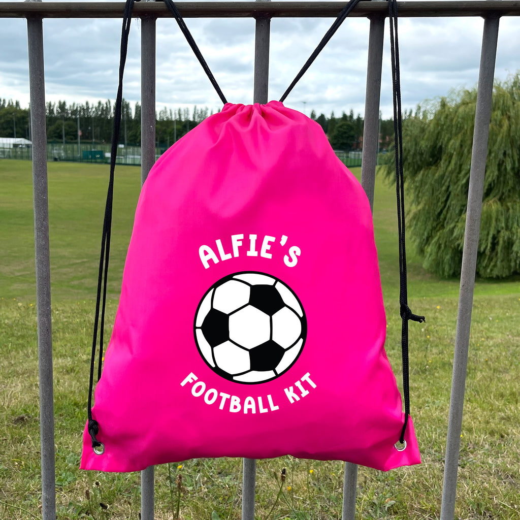 Pink drawstring bag with soccer ball design and 'Alfie's Football Kit' text, hanging on a metal fence outdoors.