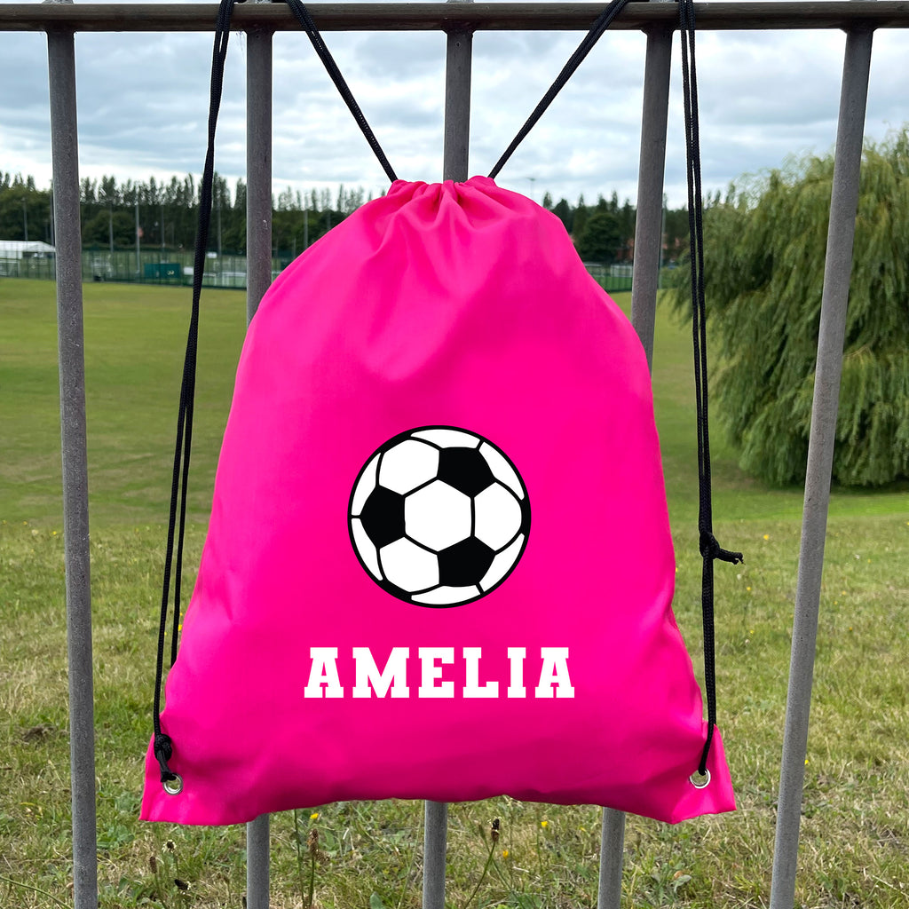 Pink drawstring bag with soccer ball design and 'AMELIA' text, hanging on a metal fence with a grassy field background.