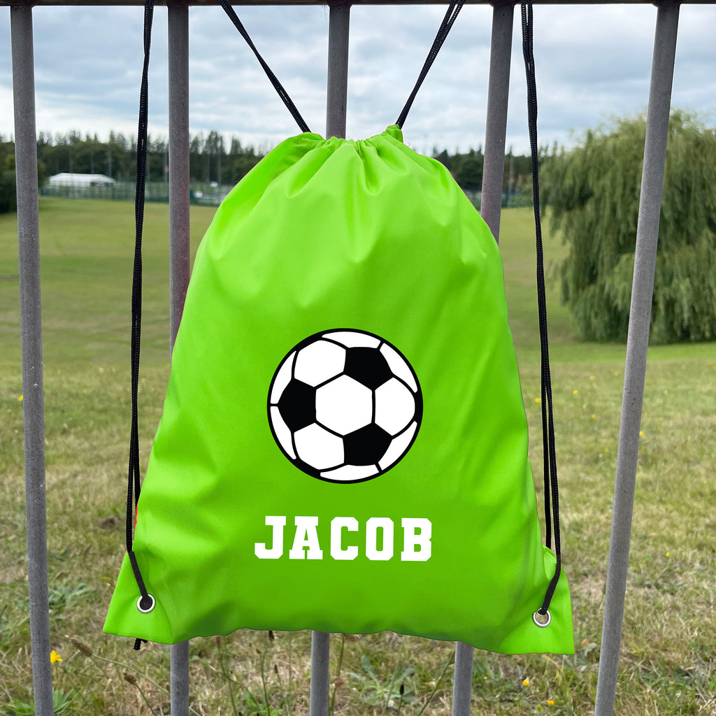 Green drawstring bag with soccer ball design and 'JACOB' text, hanging on a metal fence with a grassy field background.