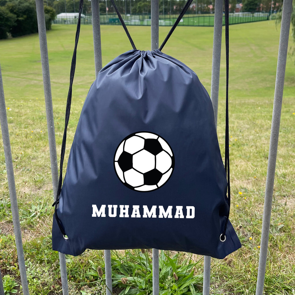 Navy drawstring bag with soccer ball design and 'MUHAMMAD' text, hanging on a metal fence with a grassy field background.