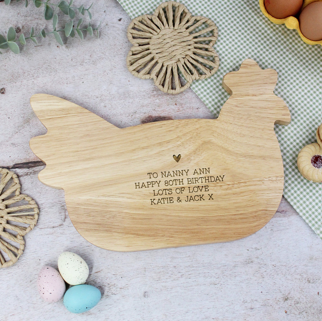 Wooden cutting board shaped like a rooster with engraved text on a table with Easter eggs and a basket.