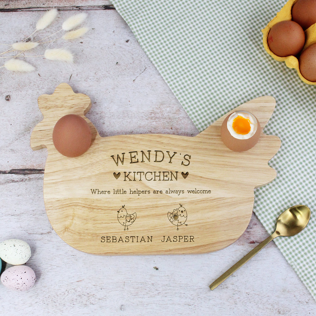 Wooden cutting board shaped like a chicken with engraved text, surrounded by eggs and a spoon on a checkered cloth.