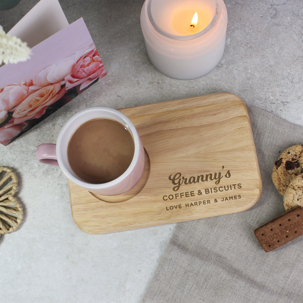 Pink mug on a wooden coaster with 'Granny's Coffee & Biscuits' text, surrounded by cookies and a candle.