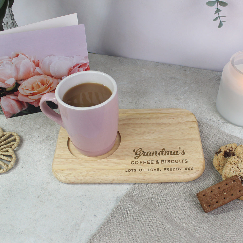 Pink mug with coffee on a wooden coaster with 'Grandma's Coffee & Biscuits' text, surrounded by cookies and a card with flowers.