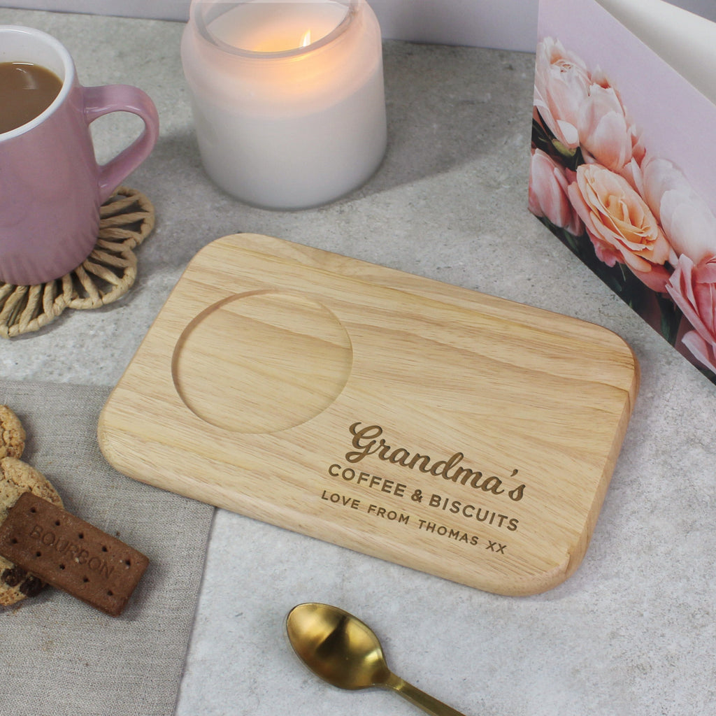 Wooden cutting board with 'Grandma's Coffee & Biscuits' engraving on a table with a cup of coffee, cookies, and a card.