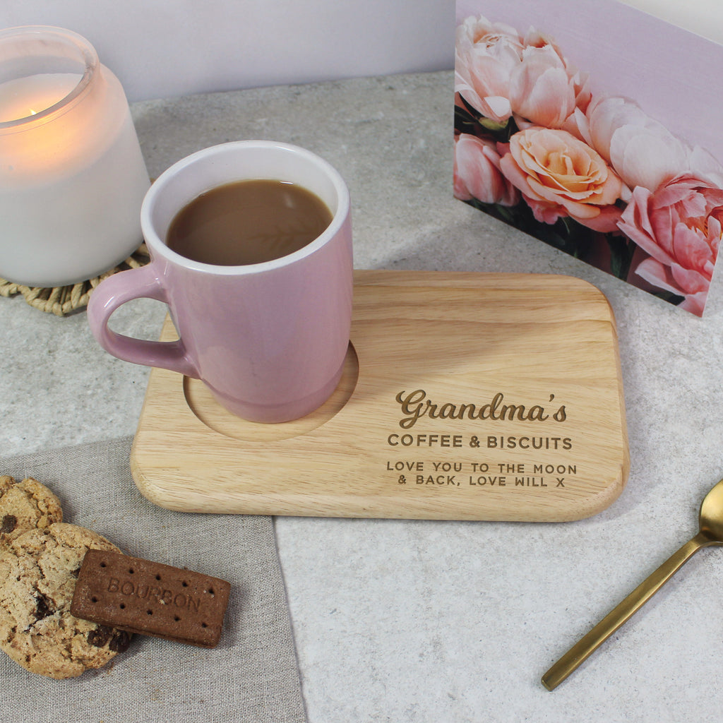 Pink mug with wooden coaster labeled 'Grandma's Coffee & Biscuits' on a table with cookies and a candle.