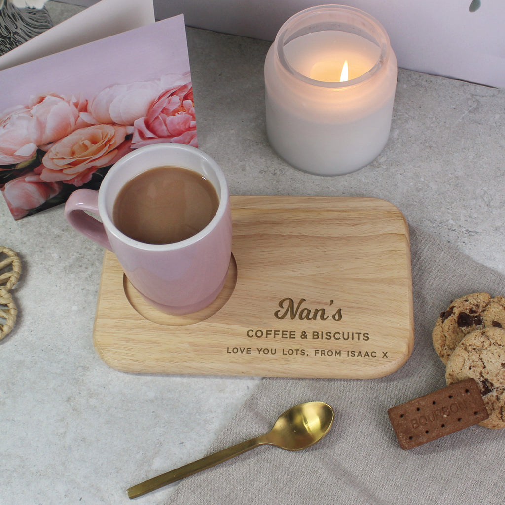 Pink mug with a wooden coaster labeled 'Nan's Coffee & Biscuits' on a table with a candle and cookies.