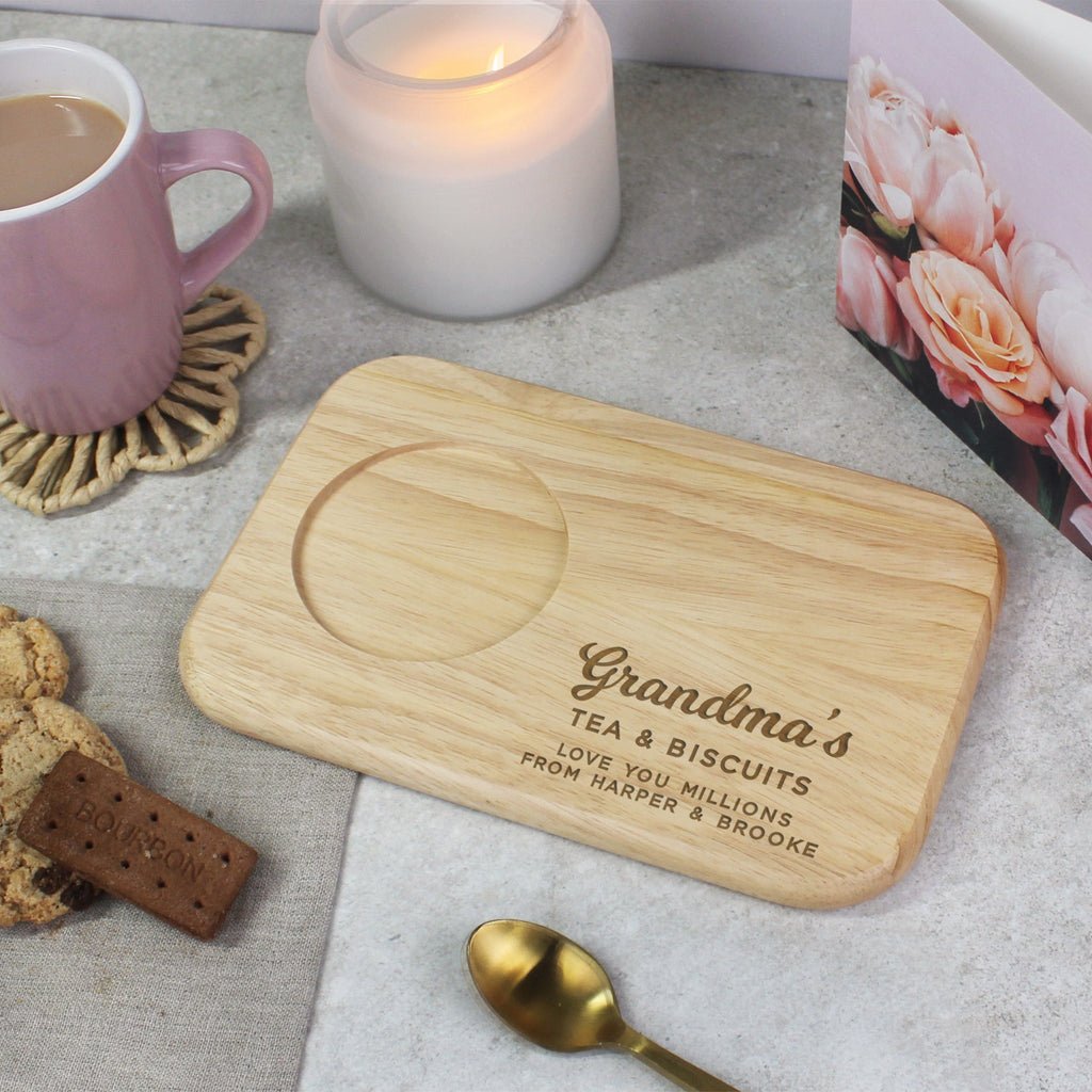 Wooden cutting board with 'Grandma's Tea & Biscuits' engraving on a table with  biscuits and a mug.