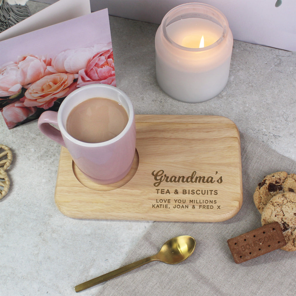 Cup of tea on a wooden coaster with 'Grandma's Tea & Biscuits' text, candle, spoon, and cookie on a light surface.