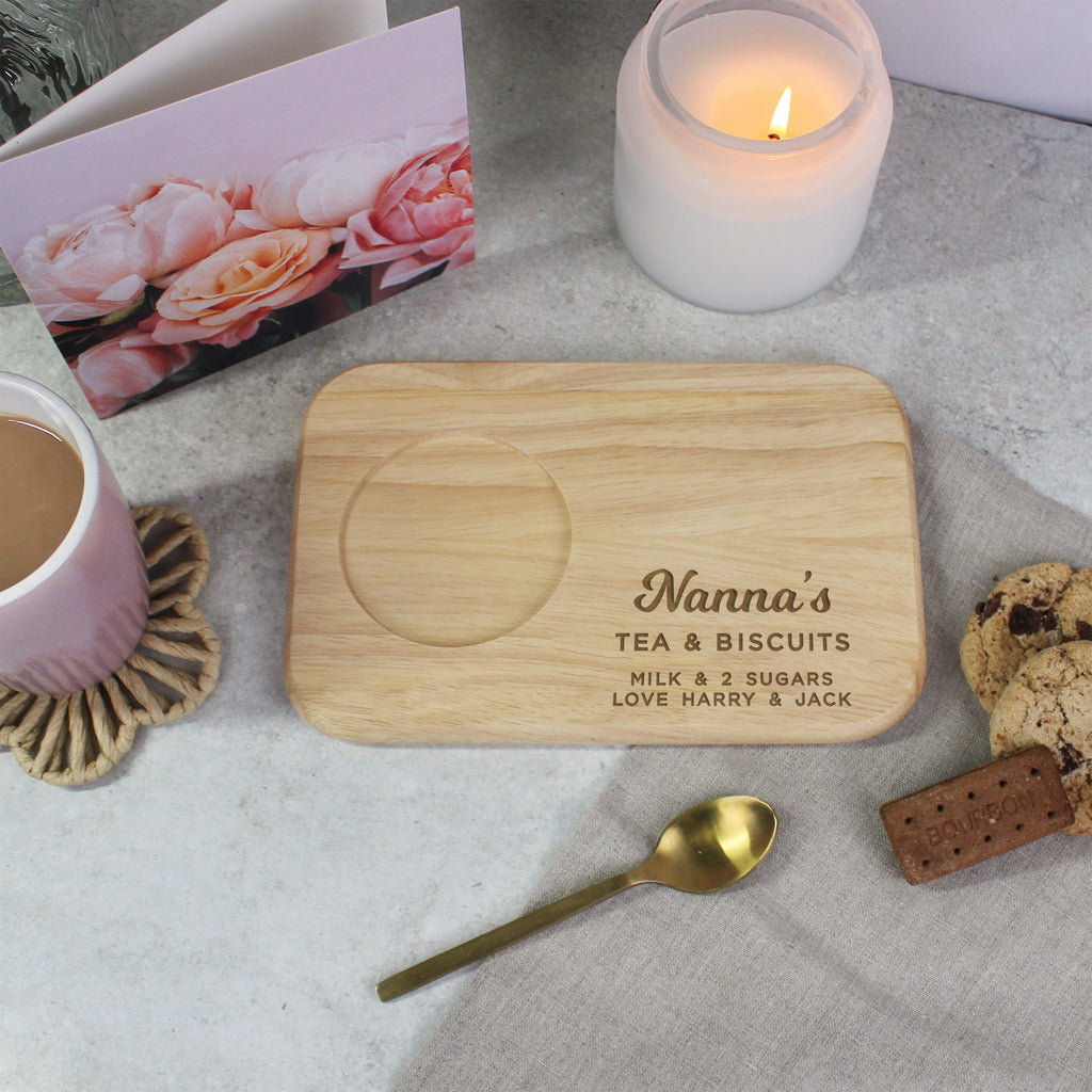 Wooden tray with engraved text on a table with a cup of tea, cookies, and a candle.