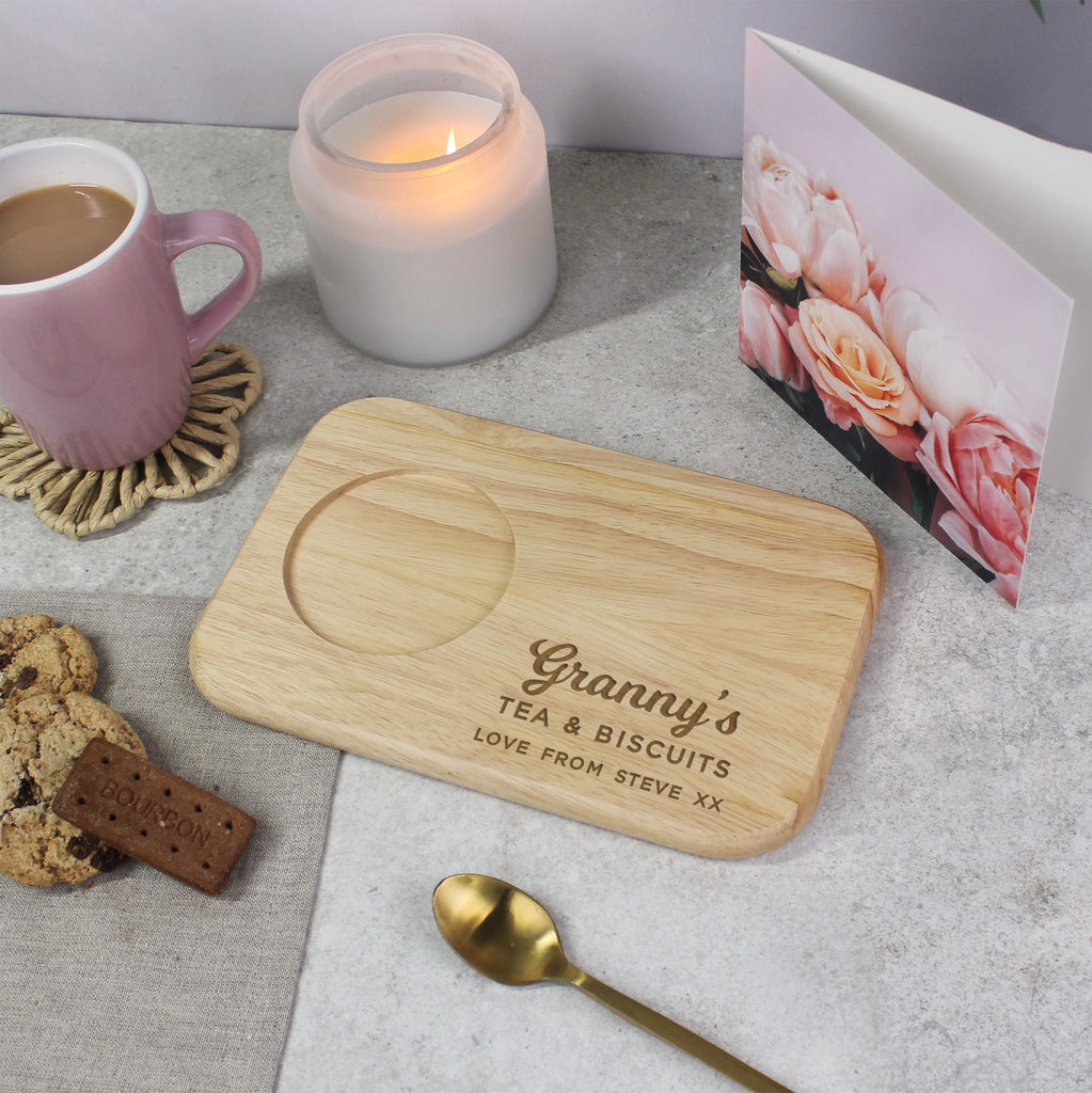 Wooden tray with 'Granny's Tea & Biscuits' engraving on a table with a mug, candle, and card.