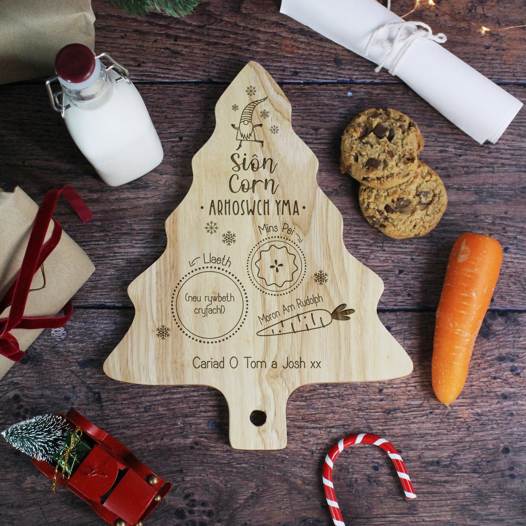 Wooden Christmas tree-shaped cutting board with engraved text on a wooden surface with cookies and a carrot.