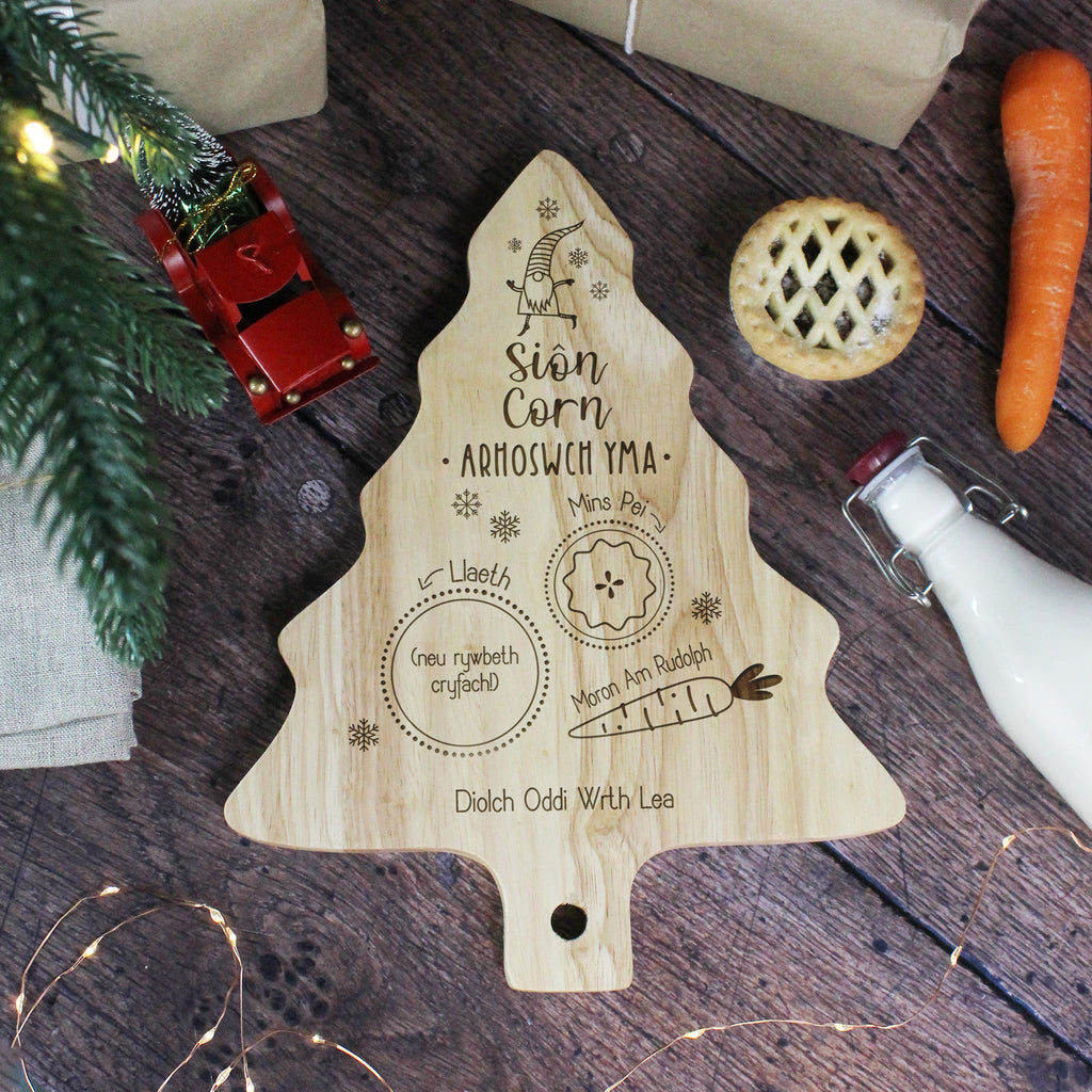 Wooden tree-shaped cutting board with engraved text on a wooden surface with Christmas decorations.