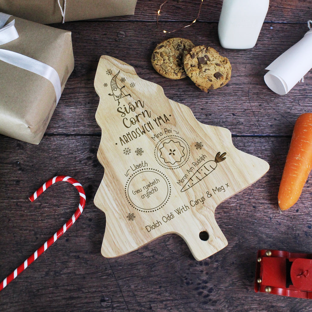 Wooden Christmas tree-shaped cutting board with engraved text on a wooden surface with cookies and candy canes.