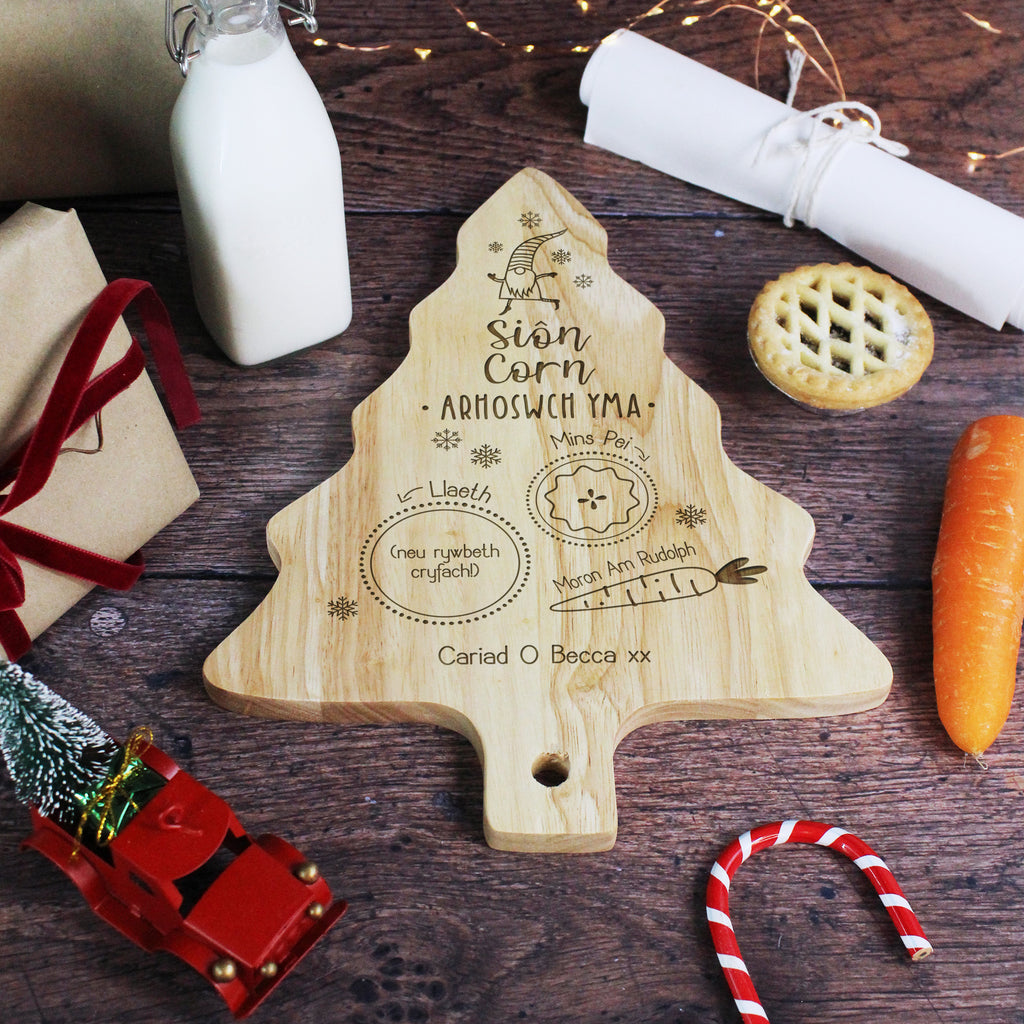 Wooden Christmas tree-shaped cutting board with engraved text on a festive wooden table.