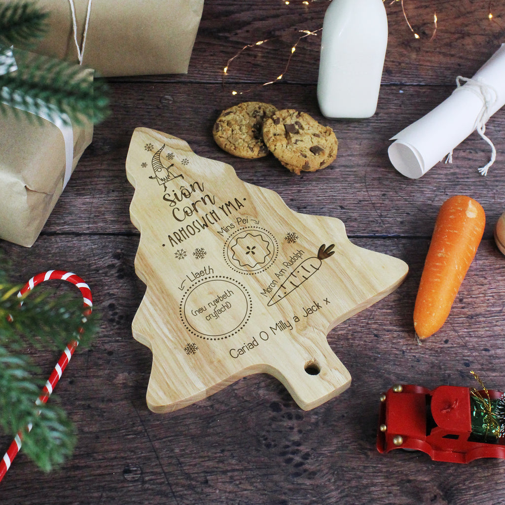 Wooden Christmas tree-shaped cutting board with engraved text on a wooden surface with cookies and a carrot.
