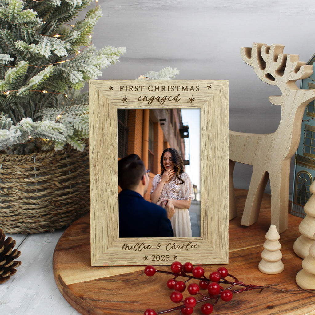 Wooden photo frame with 'First Christmas engaged' engraving, placed on a table with festive decorations.