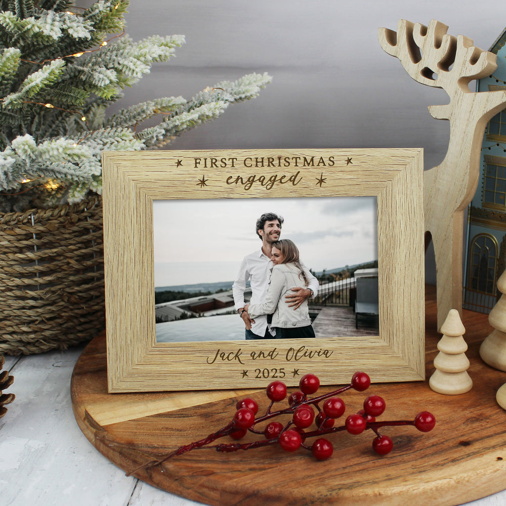 Wooden photo frame with 'First Christmas engaged' text, placed on a wooden surface with festive decorations.