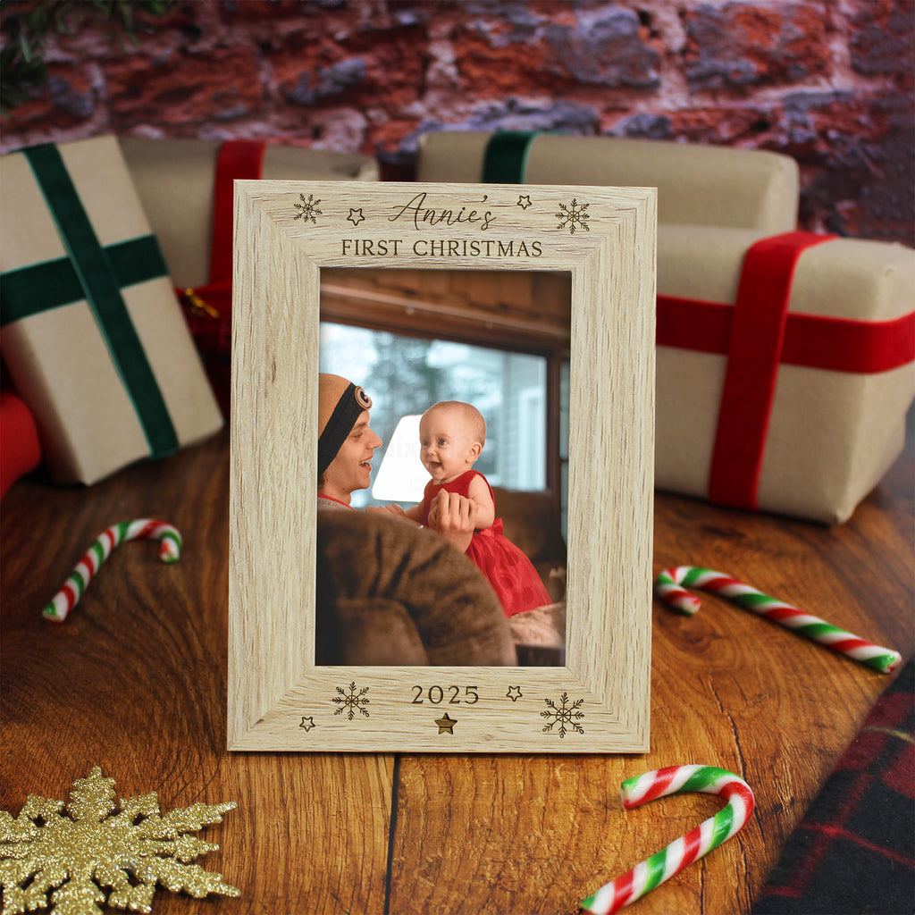Wooden photo frame with 'Annie's First Christmas 2025' text, surrounded by Christmas decorations on a wooden surface.