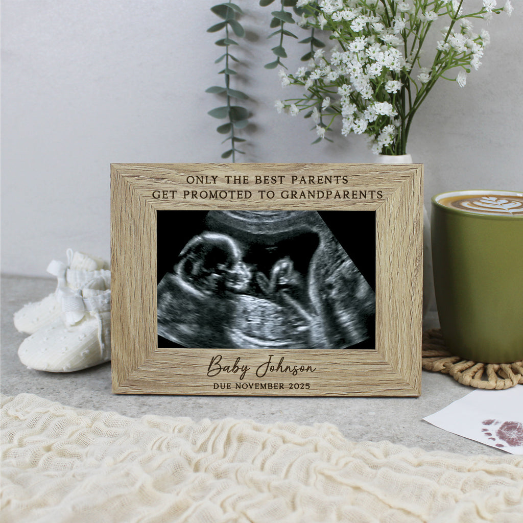 Wooden frame with ultrasound photo and text 'Only the best parents get promoted to grandparents' on a surface with a cup and flowers.