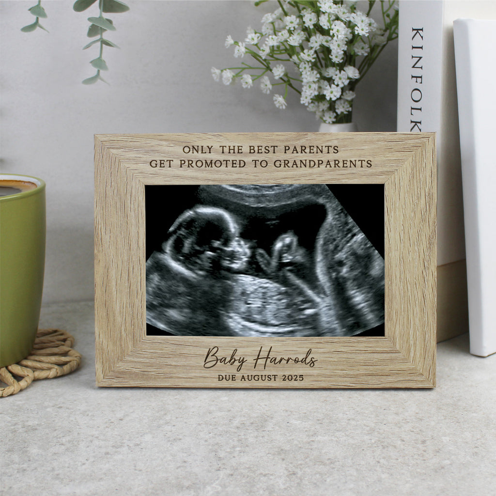Wooden picture frame with ultrasound photo and engraved text on a surface with a cup and flowers in the background.
