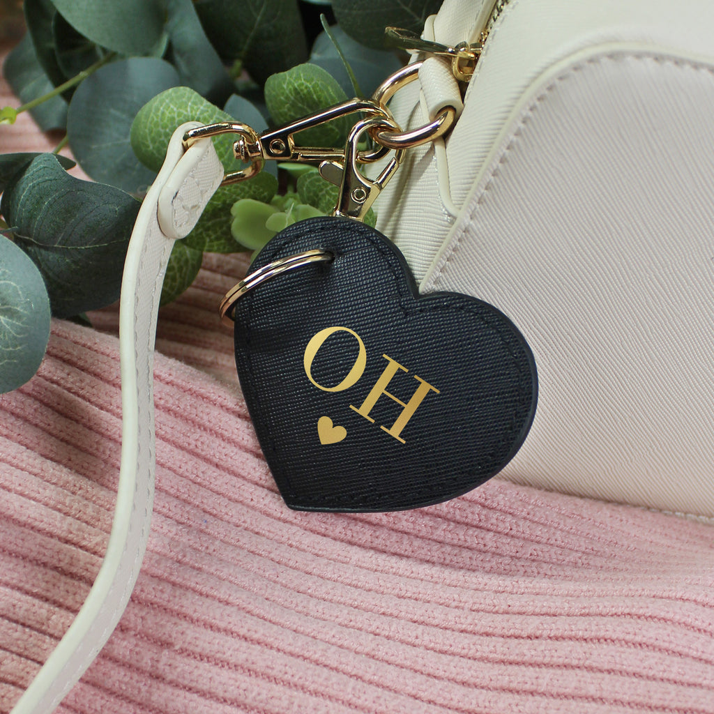Black heart-shaped keychain with 'OH' initials on a pink surface with green leaves in the background