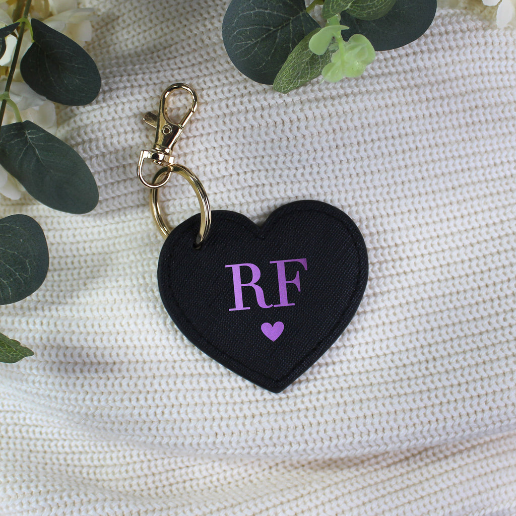 Black heart-shaped keychain with 'RF' and a purple heart on a textured surface with green leaves.