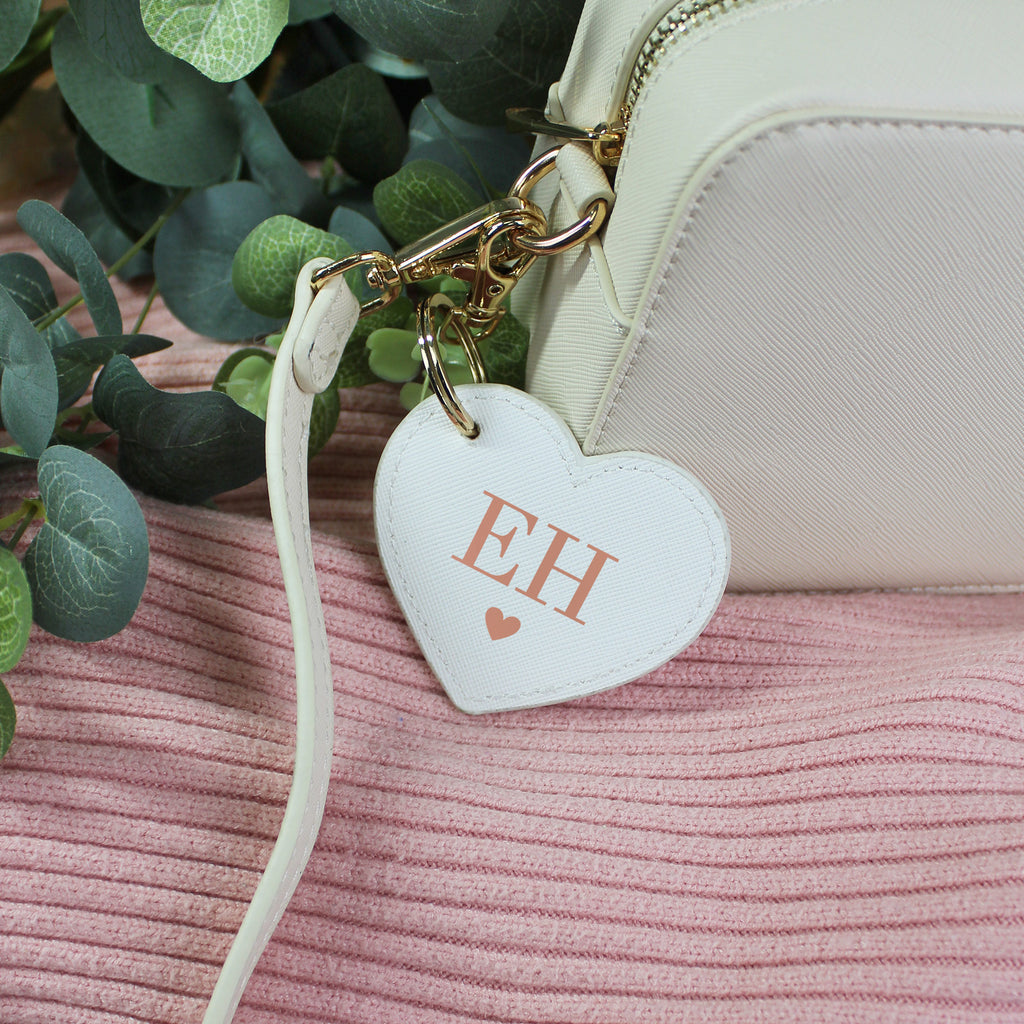 Heart-shaped keychain with 'EH' initials attached to a white bag on a pink surface with green leaves in the background.
