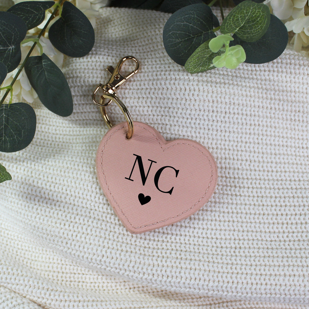 Pink heart-shaped keychain with 'NC' and a black heart on a textured white surface with green leaves.