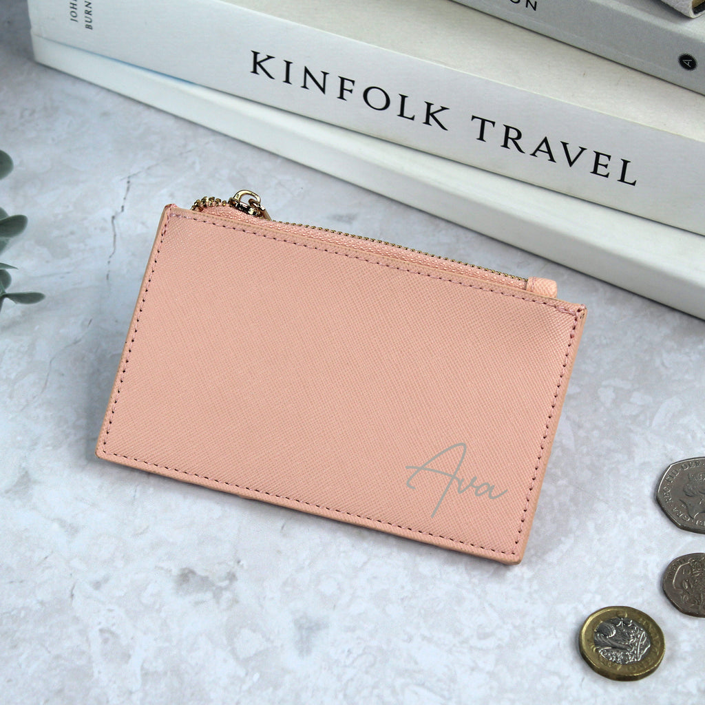 Pink wallet with 'Ava' branding on a light surface with coins and books in the background