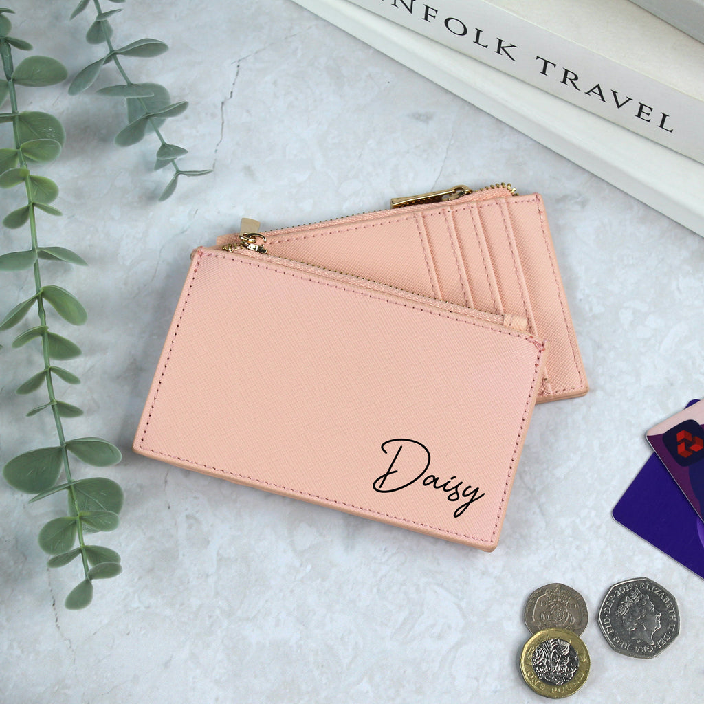 Pink leather wallet with 'Daisy' branding on a light gray surface with coins and a plant.