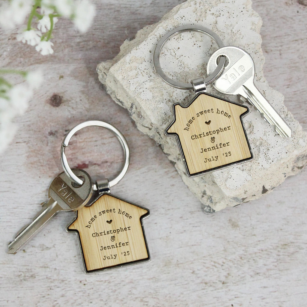 Two keychains with engraved house-shaped tags on a stone surface with white flowers.